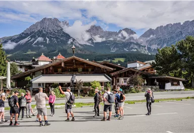 A group of people stands in front of a rustic building in the Alps. The impressive mountains are visible in the background, surrounded by clouds.
