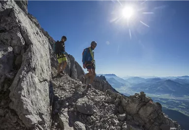 Two hikers are walking along a narrow path on the mountain. In the background, there is an impressive mountain landscape against a clear blue sky.
