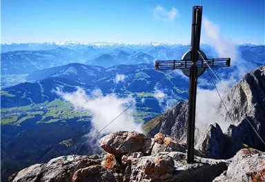An impressive mountain panorama with a cross on a summit. In the background, majestic mountains and a clear sky can be seen.