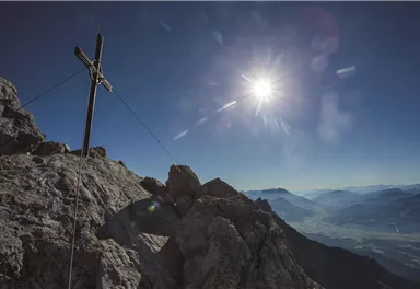 A summit with a cross and radiant sunshine. The surrounding mountains are clear and visible in the background.