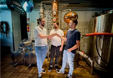 Three men are standing in a distillery holding glasses in their hands. In the background, shiny copper and stainless steel equipment can be seen.