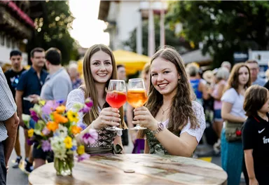 Two young women toast with colorful drinks. In the background, people and market stalls can be seen, creating a festive atmosphere.