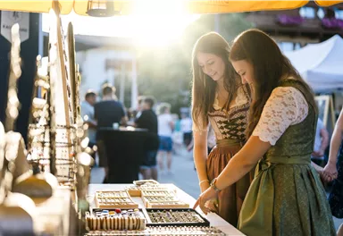 Two women in traditional clothing are looking at jewelry in a market. In the background, sunlight is visible, creating a cozy atmosphere.
