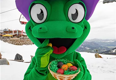 Ein fröhliches Maskottchen in einem grünen Anzug mit lila Hut steht im Schnee und hält einen Korb mit bunten Eiern. Im Hintergrund sind Berglandschaften und Skilifte sichtbar.