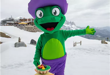 Ein grünes Maskottchen in einem lila Hut steht im Schnee und lächelt. Es hält eine Schale mit Essen in der Hand und zeigt mit der anderen Hand auf die Umgebung.