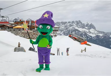 Ein grüner Zauberer in einem bunten Kostüm steht im Schnee vor einer Berglandschaft. Im Hintergrund sind ein Skilift und eine Berghütte zu sehen.