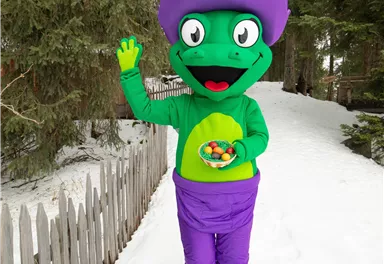Ein farbenfroher Maskottchen steht im Schnee und winkt. Es trägt einen lila Hut und hält eine Schüssel mit bunten Süßigkeiten.