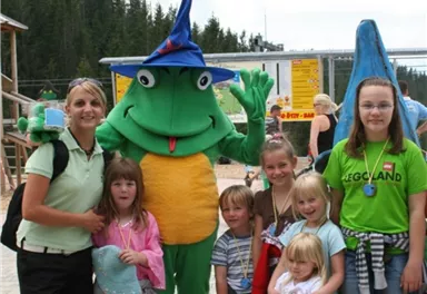 Eine Gruppe von fröhlichen Kindern und einer Frau posiert mit einem großen, grünen Maskottchen. Im Hintergrund sind Bäume und Spielgeräte zu sehen.