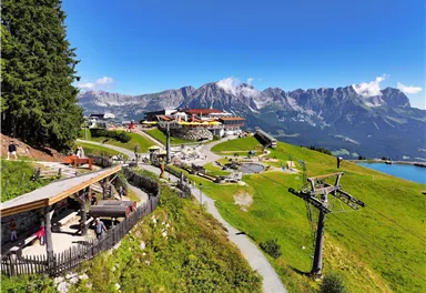 A picturesque mountain landscape with a restaurant and views of the surrounding mountains. In the foreground, hiking trails and a cable car can be seen.