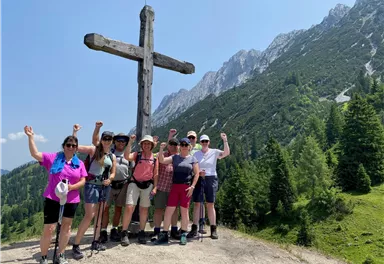 Eine Gruppe von Wanderern steht vor einem großen Holzkreuz in den Bergen. Sie lächeln und posieren fröhlich auf einem sonnigen Tag.
