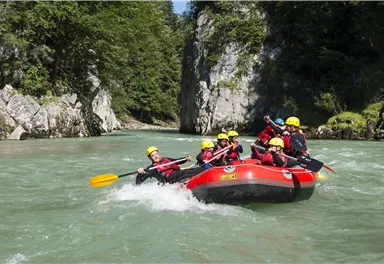 A group of people paddles in a red raft on a clear river. Surrounded by green trees and rocks, the scenery provides an adventurous experience in nature.