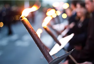 Eine Gruppe von Menschen hält Fackeln in der Dämmerung. Die Flammen leuchten in der Dunkelheit und schaffen eine feierliche Atmosphäre.