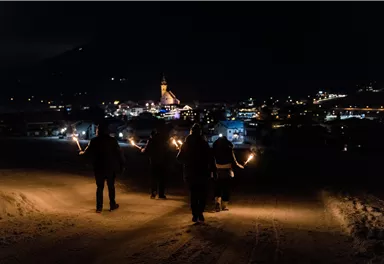 A group of people with torches walks through a snow-covered landscape at night. In the background, a city with a church is glowing.