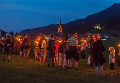 Eine Gruppe von Menschen mit Fackeln geht durch eine Wiese bei Dämmerung. Im Hintergrund ist eine Kirche und eine malerische Landschaft zu sehen.