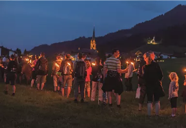 A group of people with torches gathers in nature at twilight. In the background, a church and rolling hills can be seen.