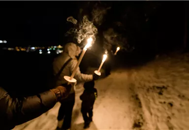 Eine Gruppe von Menschen geht nachts mit Fackeln durch den Schnee. Die Umgebung ist beleuchtet von den Lichtern der Stadt in der Ferne.