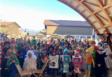 Eine große Gruppe von Kindern in bunten Kostümen versammelt sich unter einem Holzdach. Im Hintergrund sind Berge und ein klarer Himmel zu sehen.