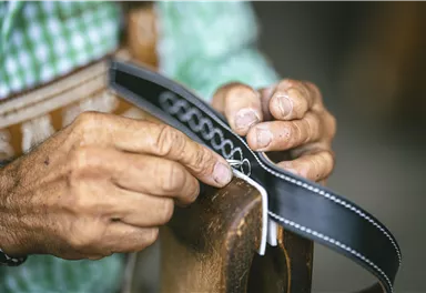 Ein Handwerker arbeitet sorgfältig an einem schwarzen Lederstück. Die Hände sind gekennzeichnet von der Handarbeit und zeigen viel Geschick.