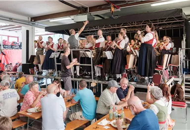 Eine Blaskapelle spielt auf einer Bühne, während das Publikum an Tischen sitzt. Die Menschen genießen das Essen und die Musik in einer geselligen Atmosphäre.