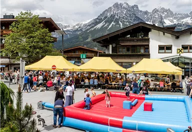 Eine lebhafte Veranstaltung im Freien mit gelben Zelten und vielen Besuchern. Im Vordergrund gibt es eine aufblasbare Spielarena und eine malerische Bergkulisse.