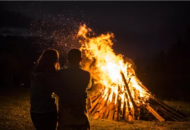 Ein Lagerfeuer in der Nacht strahlt warmes Licht aus. Zwei Personen stehen nah beieinander und schauen dem Feuer zu.