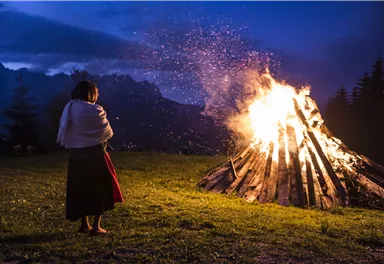 A large campfire is burning in the twilight, sparks are flying into the air. A person is standing in the foreground, looking at the fire.