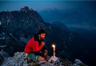 A man sits on a mountain peak at night and lights a candle. In the background, impressive mountains and the twilight light can be seen.