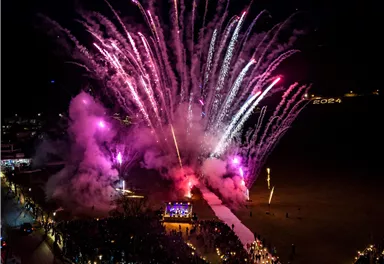 A spectacular firework illuminates the night sky with vibrant colors. Many people are celebrating and enjoying the festive atmosphere on the beach.