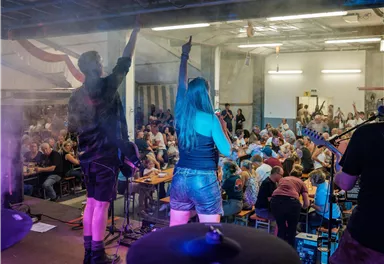 Eine Band spielt auf einer Bühne vor einem begeisterten Publikum. Die Zuschauer heben ihre Hände und feiern die Musik.