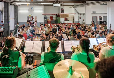 Eine große Veranstaltung mit vielen Menschen, die an Tischen sitzen. Im Vordergrund spielen Musiker in traditioneller Kleidung.