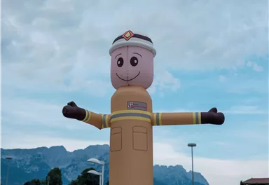 Ein aufblasbarer Feuerwehrmann mit einem freundlichen Gesicht steht mit offenen Armen. Im Hintergrund sind Berge und ein bewölkter Himmel zu sehen.