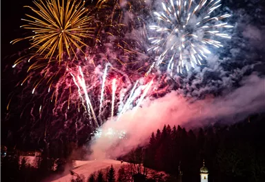 A spectacular fireworks display lights up the night sky above a snowy landscape. In the foreground stands a small church surrounded by trees.