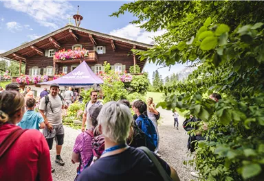 A group of people stands in front of a traditional wooden house with blooming windows. In the background, there is a green landscape that highlights the beautiful day.