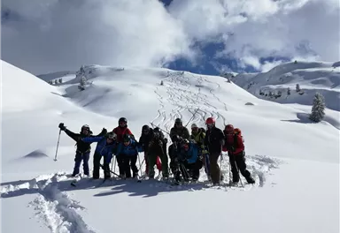 Eine Gruppe von Skifahrern steht lächelnd im frischen Schnee. Im Hintergrund sind verschneite Hügel und eine klare Himmelslandschaft zu sehen.