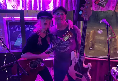 Zwei Musiker stehen zusammen auf einer Bühne. Sie halten Instrumente und lächeln in die Kamera, während die Umgebung in bunten Lichtern beleuchtet ist.