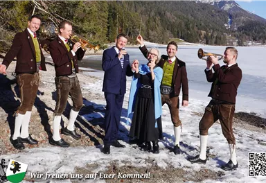 A group of six people is standing by a frozen lake. Some are wearing traditional clothing and playing musical instruments, while others are cheerfully posing.