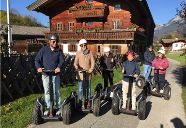 A group of people is riding Segways on a narrow path. In the background, a traditional wooden house with flower boxes can be seen.