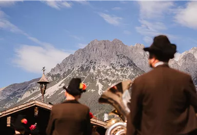 Zwei Männer in traditionellen Trachten stehen im Vordergrund und blicken auf majestätische Berge. Der Himmel ist blau mit einigen Wolken.