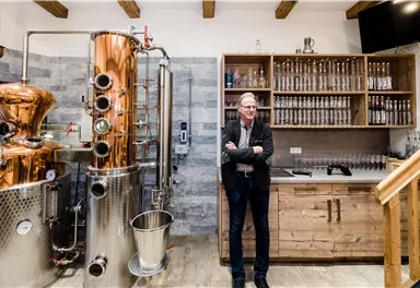 A modern distillery with a copper kettle and wooden shelves. A man stands relaxed next to the equipment.