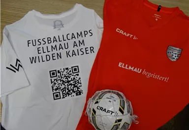 Ein rotes Fußballtrikot und ein weißes T-Shirt liegen auf einem Tisch. Daneben liegt ein Fußball mit dem Schriftzug "CRAFT".