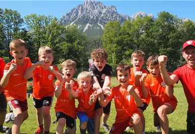 Eine Gruppe fröhlicher Kinder in orangefarbener Sportkleidung posiert auf einem grünen Feld. Im Hintergrund sind hohe Berge und ein blauer Himmel zu sehen.