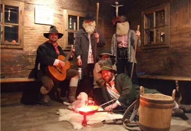Eine Gruppe von fünf Personen in historischer Kleidung steht in einem rustikalen Raum. Sie posieren um ein Feuer in einem Holzfass und haben verschiedene Requisiten.