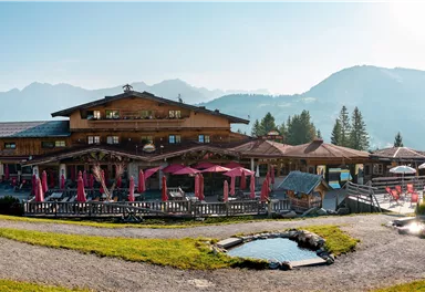 Ein malerisches Alpenhaus mit einer einladenden Terrasse und Blick auf die Berge. Die Umgebung ist grün und sonnig, ideal für einen entspannten Aufenthalt.
