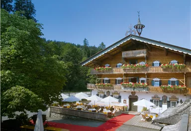 A traditional alpine hotel with a wooden facade and blooming balconies. On the terrace, there are tables and sun umbrellas amidst green trees.