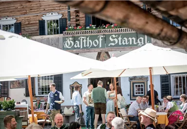 A lively terrace of a guesthouse with guests under sun umbrellas. In the background, the sign "Gasthof Stangl" is visible.
