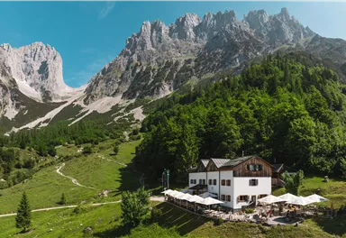 Eine malerische Berghütte umgeben von hohen Bergen und grünen Wäldern. Auf der Terrasse stehen Tische mit Sonnenschirmen.