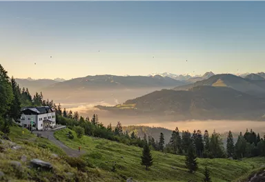 A picturesque mountain landscape with green meadows and dense forests. In the foreground stands a cozy house, surrounded by gentle hills and the expanse of the valley.