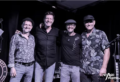 Eine Rockband posiert auf dem Dach eines Flugzeugs. Im Hintergrund sieht man einen Flugturm und einen bewölkten Himmel.