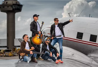 Eine Band posiert auf einem Flugzeugdach, umgeben von Wolken und einem Flughafengebäude. Die Musiker halten Gitarren und tragen lässige Kleidung.