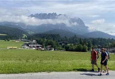 Two people are walking on a path in a green landscape. In the background, majestic mountains rise up with clouds.